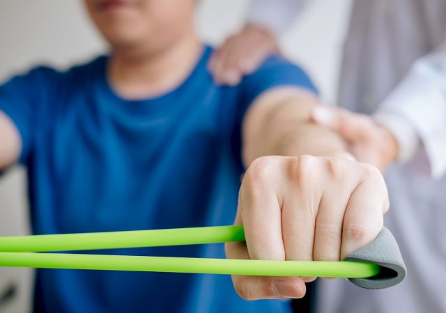 Doctor physiotherapist assisting a male patient while giving exercising treatment on stretching his arm with exercise band in the clinic, Rehabilitation physiotherapy concept.