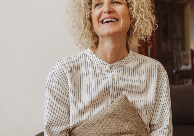 Beautiful happy smiling relaxed middle aged woman relaxing on sofa at home. Smiling stylish middle aged 50s lady sitting on couch holding a pillow in modern living room.